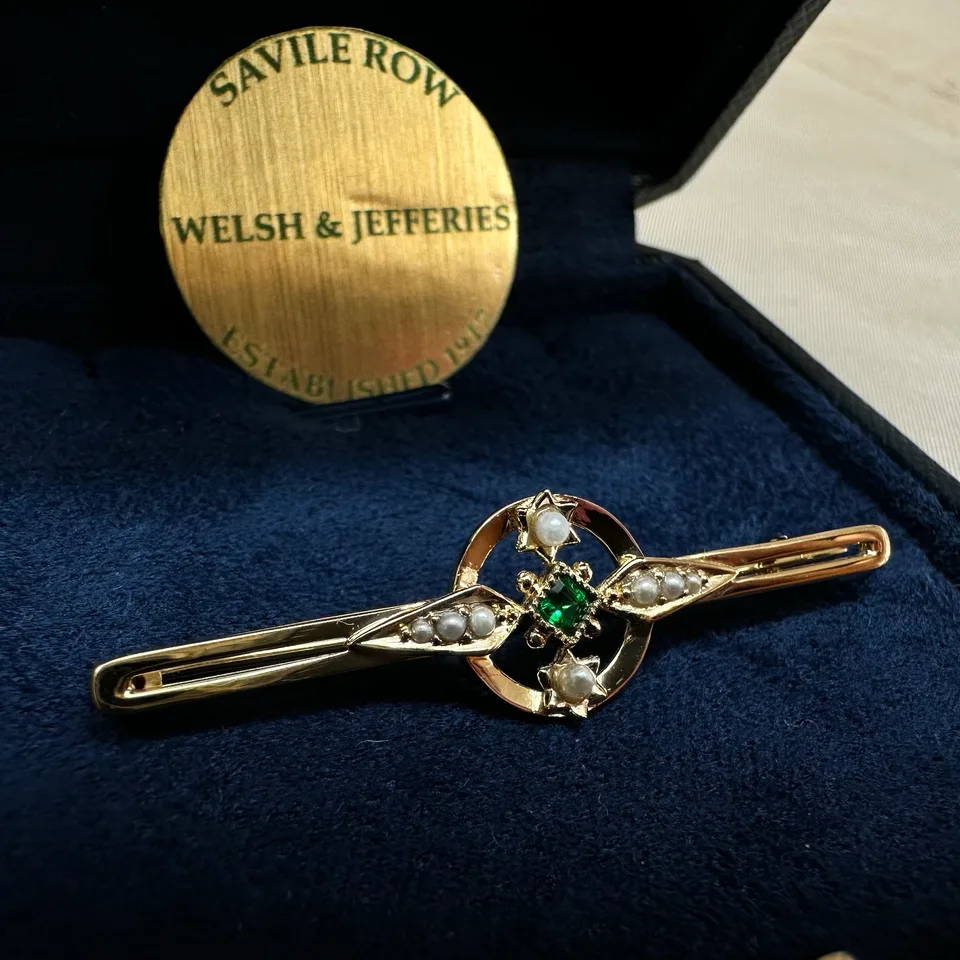 Emerald and seed-pearl gold tie bar presented on navy velvet beneath a Welsh & Jefferies Savile Row plaque.