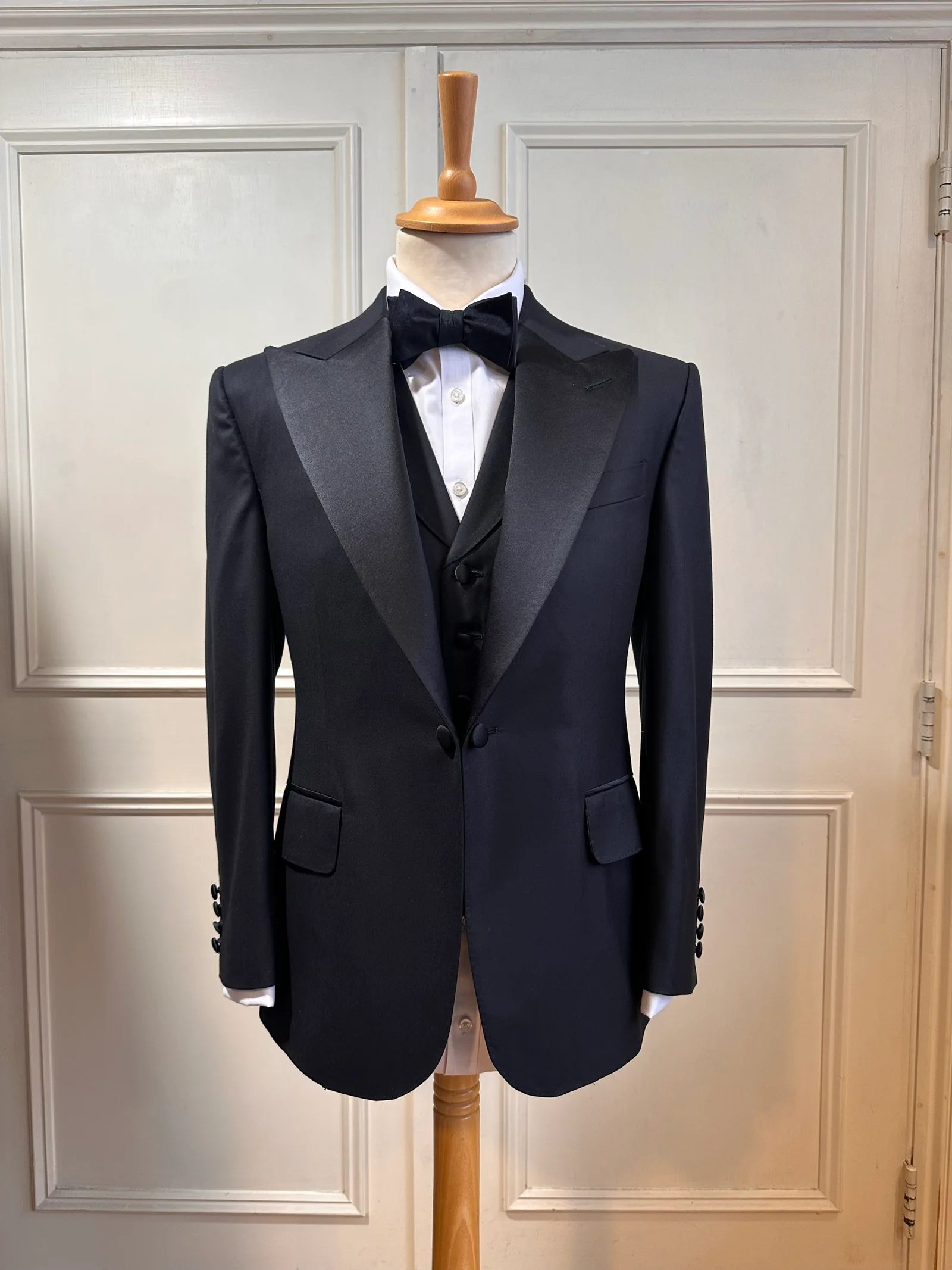 Front mannequin view of the black satin peak-lapel tuxedo, tailored with broad satin peak lapels, a clean one-button front and covered buttons.