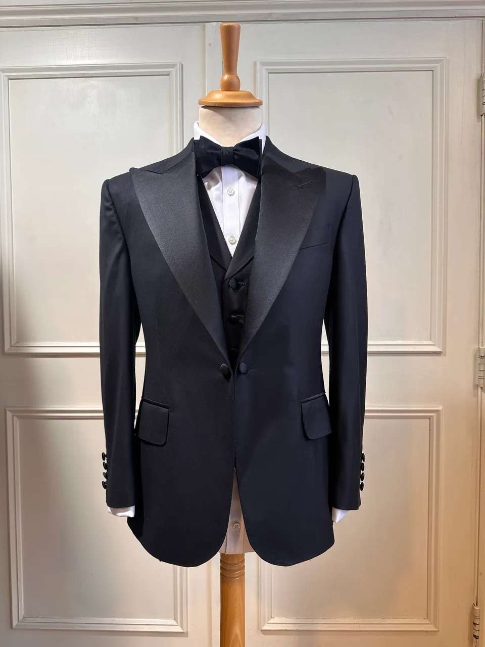 Front mannequin view of the black satin peak-lapel tuxedo, tailored with broad satin peak lapels, a clean one-button front and covered buttons.