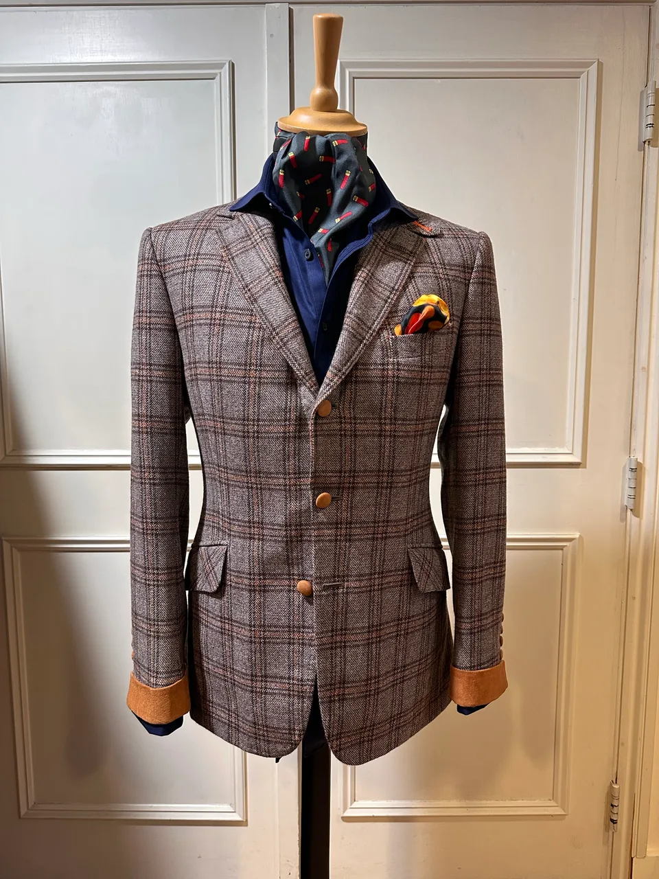 Front mannequin view of a brown Glen-check jacket with suede cuffs, showing notch lapels, three brown horn buttons, shaped waist and tan suede cuffs.