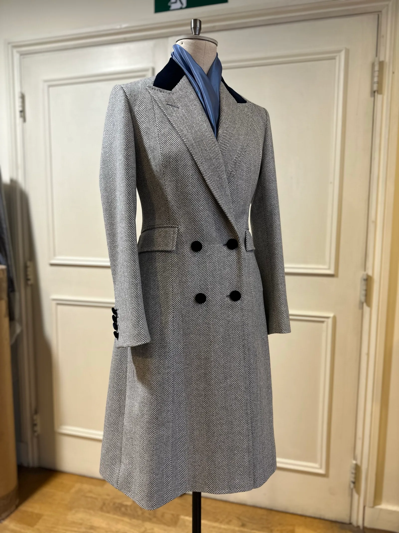 Mannequin view of a grey herringbone double-breasted overcoat, finished with broad peak lapels, a navy velvet collar and black buttons.