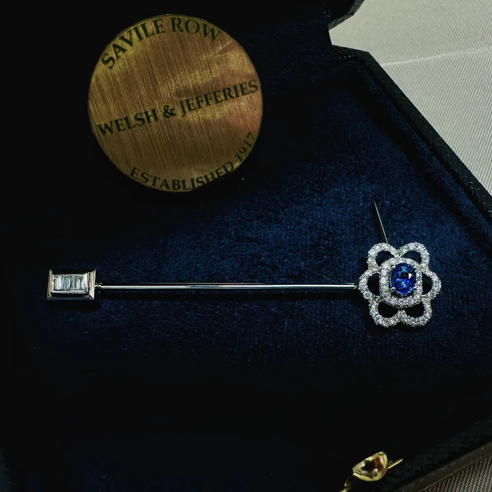 Blue sapphire stick pin encircled by airy diamond petals, with a baguette-set rectangular guard on navy velvet.