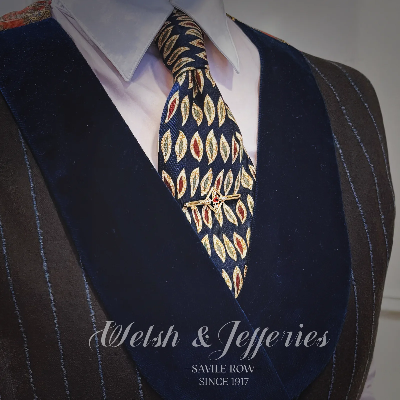 Gold compass-like tie bar centred with a round ruby and bright stone accents against navy leaf-patterned silk.