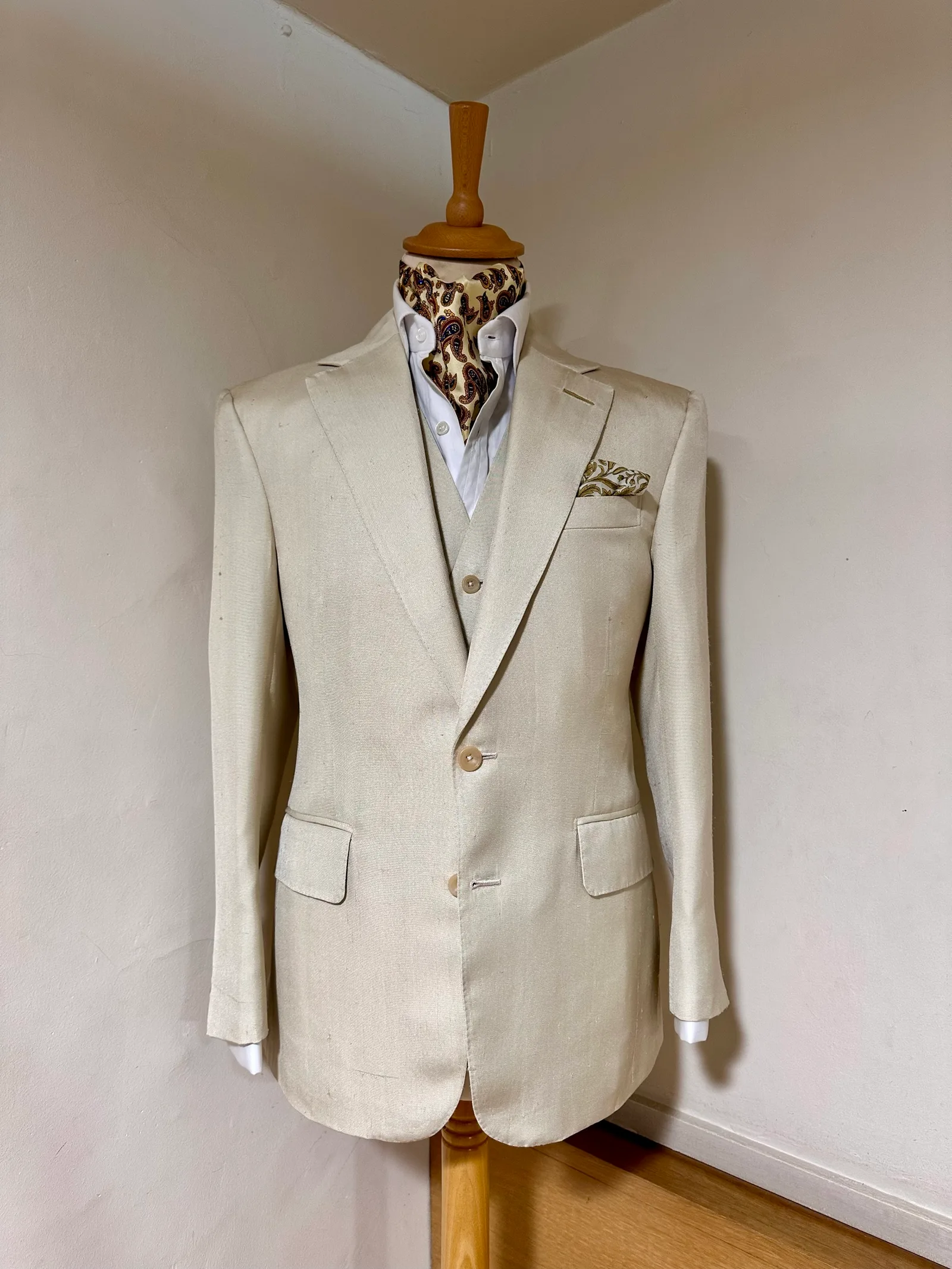 Mannequin view of an ivory summer three-piece suit, with a notch-lapel jacket, pale buttons and a matching six-button waistcoat.