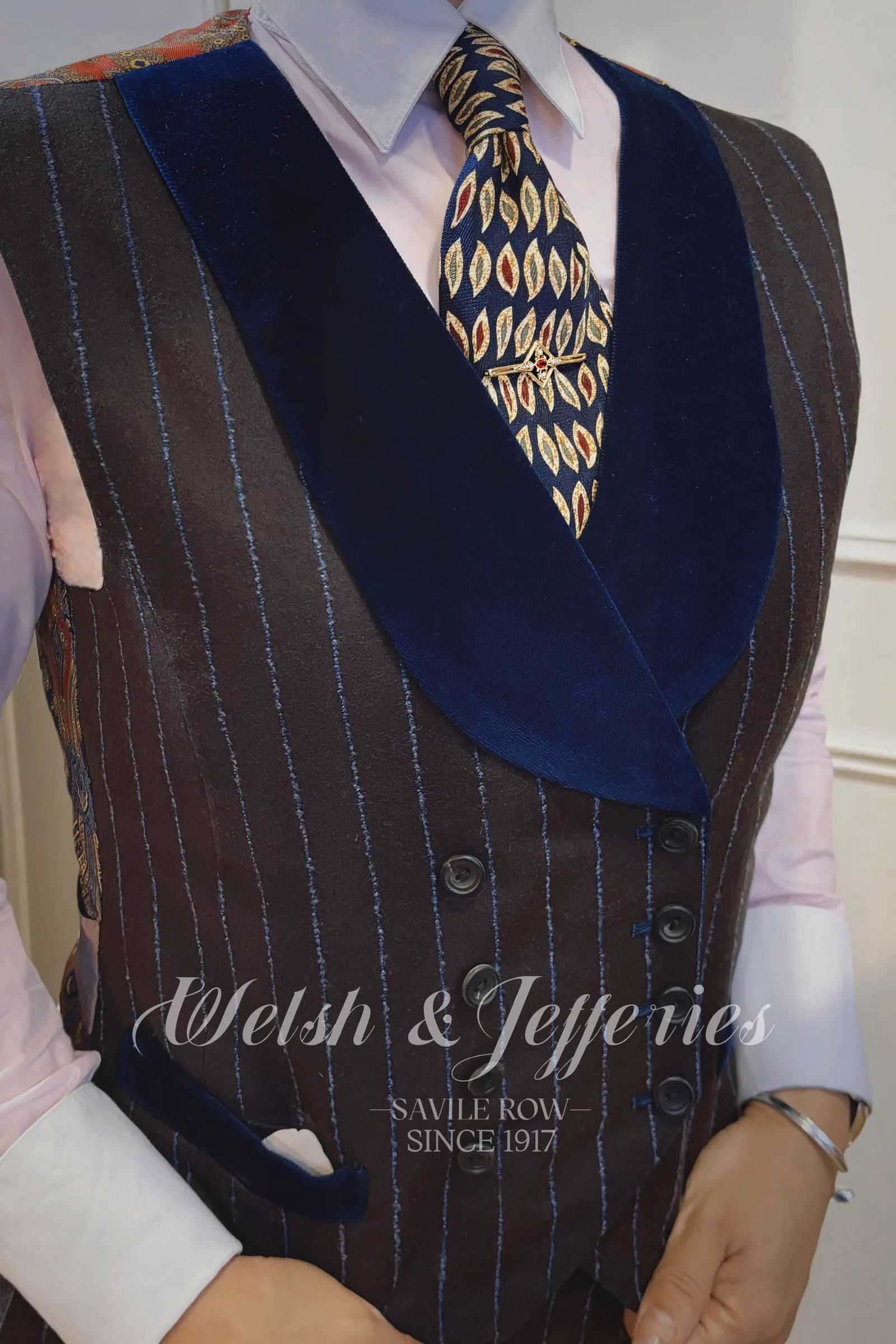 Gold geometric tie bar with a small ruby centre worn on a leaf-patterned tie and dark bespoke waistcoat.