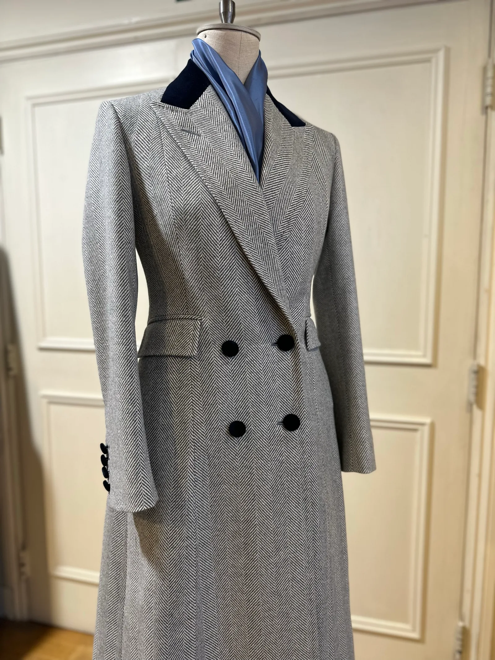 Mannequin view of a grey herringbone double-breasted overcoat, finished with broad peak lapels, a navy velvet collar and black buttons.