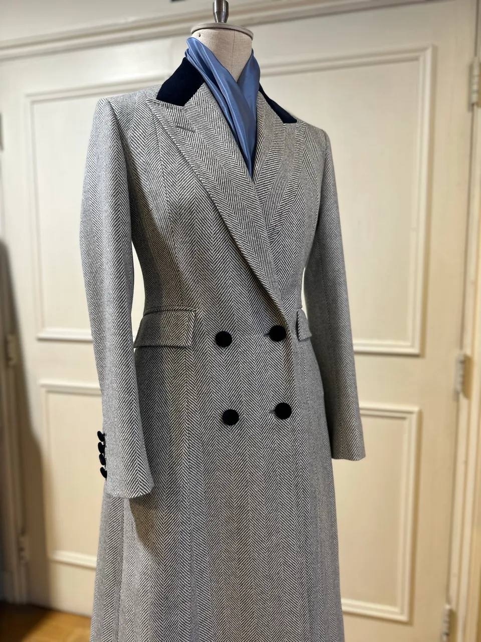 Mannequin view of a grey herringbone double-breasted overcoat, finished with broad peak lapels, a navy velvet collar and black buttons.