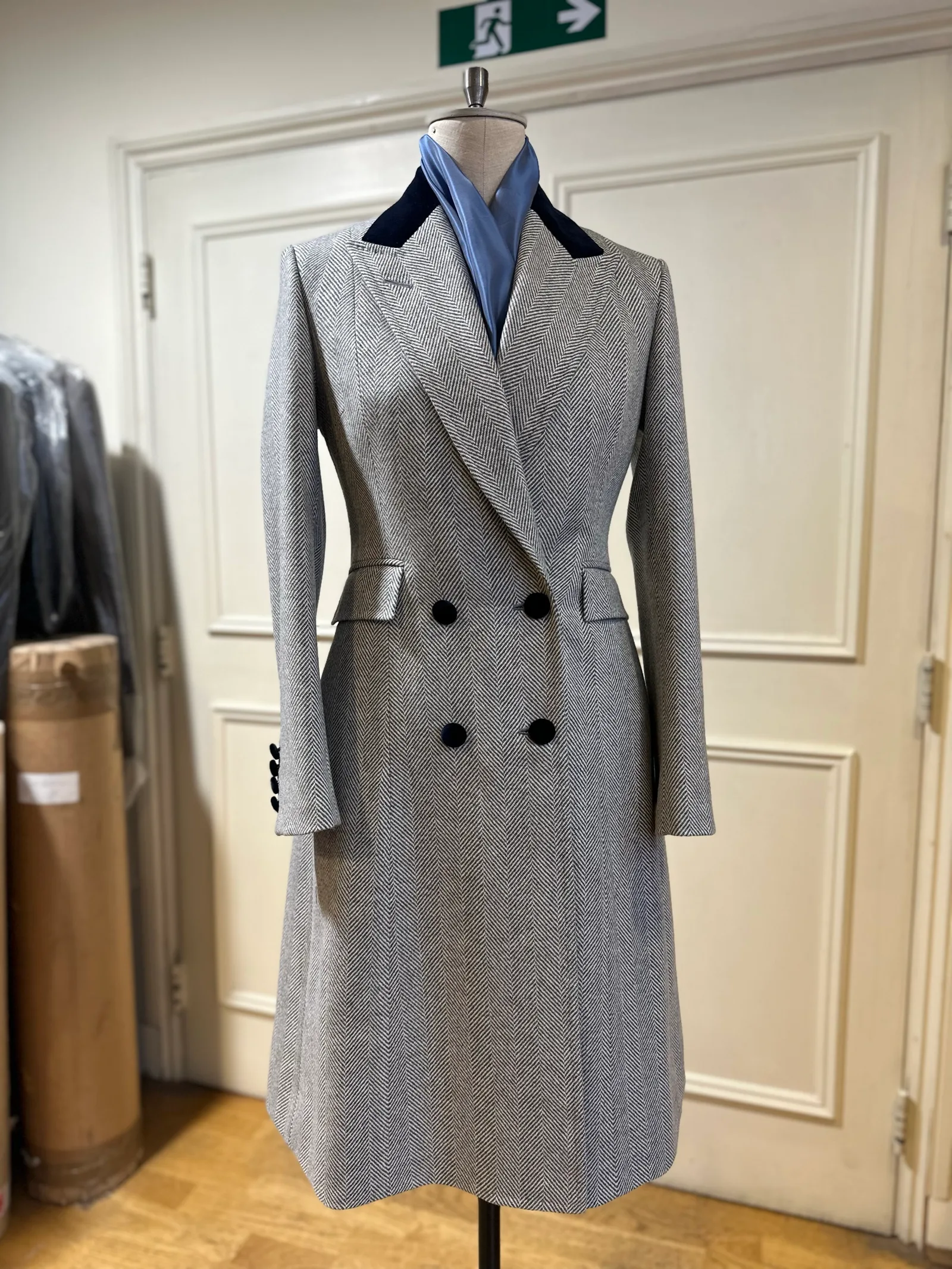 Mannequin view of a grey herringbone double-breasted overcoat, finished with broad peak lapels, a navy velvet collar and black buttons.