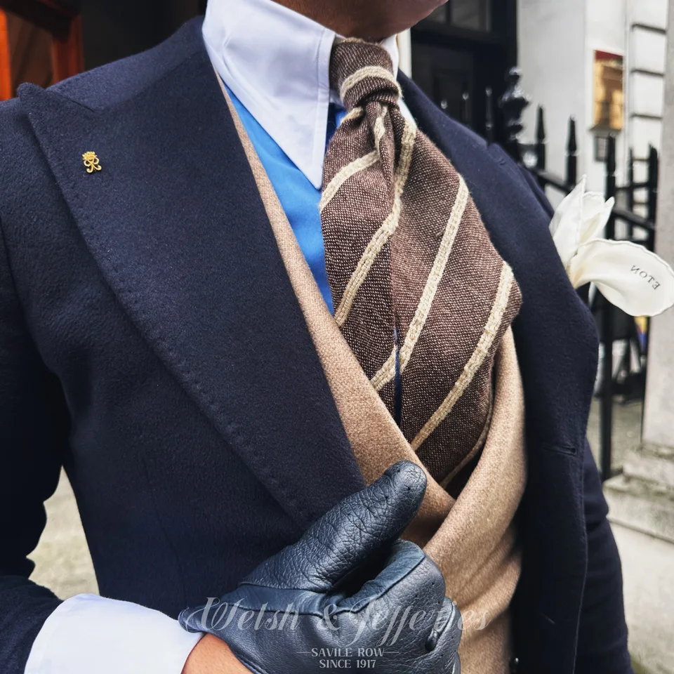 Miniature crowned gold cypher pin on a navy lapel, styled with a striped wool tie and cream boutonniere.