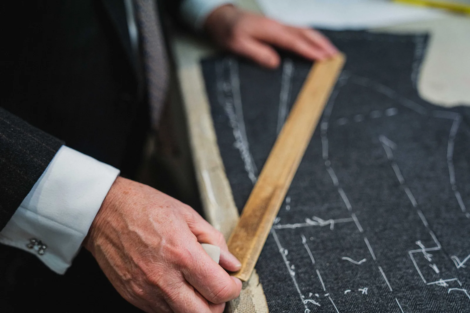 A cutter measures a hand-chalked jacket pattern across dark wool at the table.