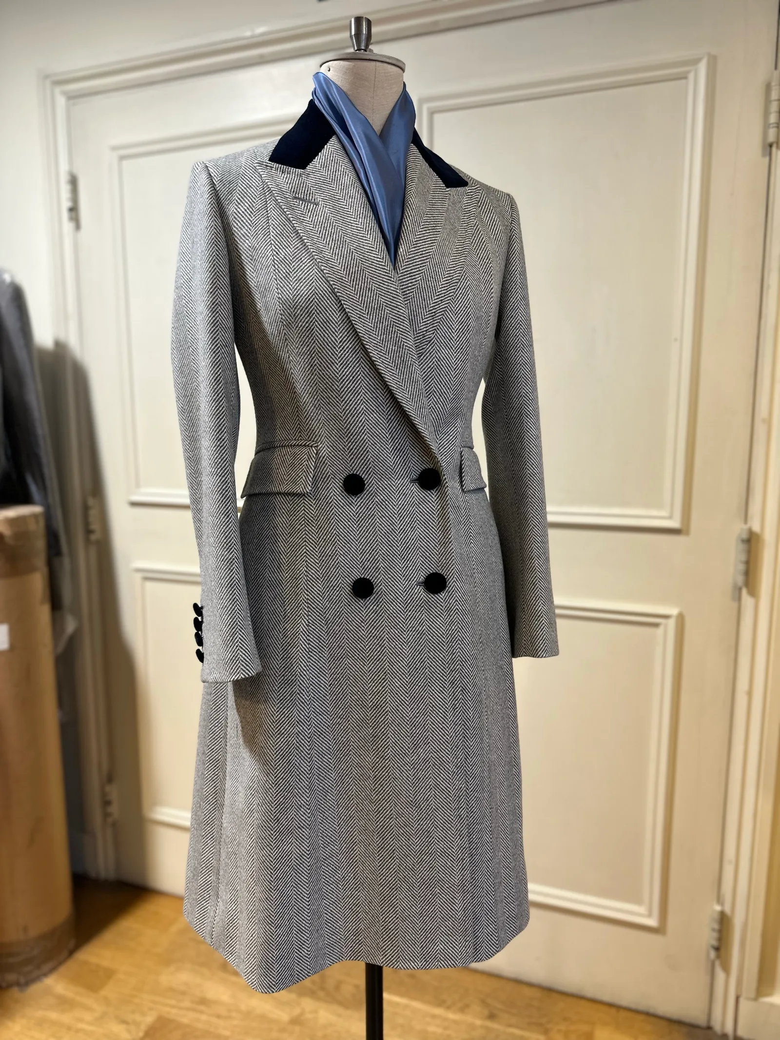 Mannequin view of a grey herringbone double-breasted overcoat, finished with broad peak lapels, a navy velvet collar and black buttons.