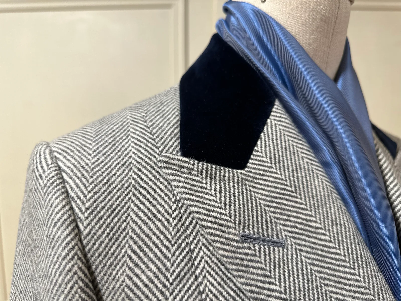 Close view of the grey herringbone overcoat, pairing a broad peak lapel with a deep navy velvet collar.